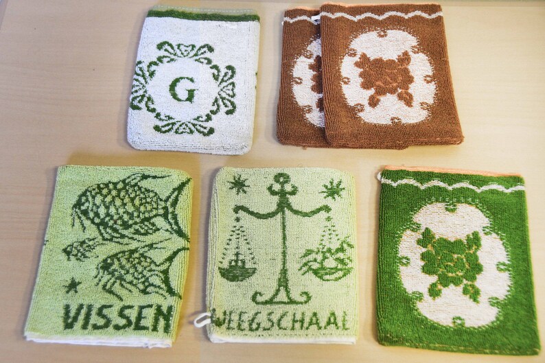 May include: Five green and white embroidered hand towels with different designs. The towels feature a letter G, a fish, a scale, and two floral designs.