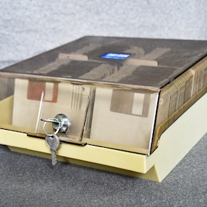 Op de afbeelding: Een beige plastic opbergdoos met een doorzichtig plastic deksel voor floppy disks. De doos heeft een slot en een sleutel.