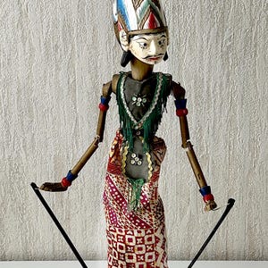 Op de afbeelding: Een Wayang Golek stokpop met een wit gezicht en geschilderde zwarte gelaatstrekken, voorzien van een hoge rood-wit-blauwe hoofdtooi. De pop heeft dunne houten armen met rode en blauwe banden, een groene kraag met franjes en een rood-witte sarong.