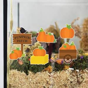May include: A decorative display featuring pumpkin patch themed stickers. The stickers include orange pumpkins, a wooden sign with the text "PUMPKIN PATCH", and a small black bird. The pumpkins are placed on a bed of wood shavings and greenery.
