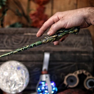 Op de afbeelding: Een groene en gouden toverstaf met ingewikkelde details, vastgehouden in een hand. De toverstaf heeft een draakachtig hoofd aan de bovenkant.