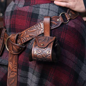 Brown floral leather coin pouch shown in hand as part of a Renaissance Faire herbalist or witch costume, made from vegetable tanned leather and suede. The embossed leaf pattern matches the bracers and belt, ideal for elven characters or nobles who need a stylish pouch for LARP or renfaire. The image shows how the pouch opens with the internal suede bag expanding for coins or trinkets, keeping the elegant fantasy aesthetic intact.