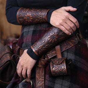 A pair of aged brown floral leather bracers is shown worn on both arms while the wearer stands in a relaxed pose with crossed arms. The renaissance faire bracer pattern displays embossed leaves and a central flower that highlight the hand dyed finish. Below, a matching floral belt and pouch complete the look, creating an elegant floral arm guard ensemble ideal as an elf cosplay accessory or larp bracer set. Perfect for fantasy themed costumes and woodland characters. Fae costume, ren faire druid