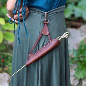 May include: A brown leather sword sheath with a decorative brass handle is attached to a belt. The sheath is secured with rivets and a leather strap. The person is wearing a teal top and a green skirt. The text "OUTCAST PROPS" is visible.