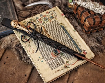 May include: A black leather-wrapped wand with a white skull at the top. The wand is lying on an old book with a brown leather cover and a fur-covered surface.