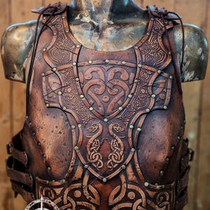 Puede incluir: Un peto de cuero marrón con intrincados diseños en relieve de nudos celtas y dragones. La armadura presenta remaches de latón y correas ajustables. El logotipo de Outcast Props es visible. Ideal para cosplay o recreaciones históricas.