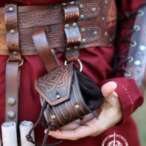 Close-up of a small decorated leather coin pouch held in hand, showing its size and texture. Handcrafted pouch with embossed Celtic geometric designs, aged brown finish, and drawstring closure. Ideal for carrying coins, dice, or keys. A compact leather accessory for Viking, Celtic, or dwarven aesthetics, suitable for LARP, Renaissance fairs, cosplay, or as a small handmade leather gift.