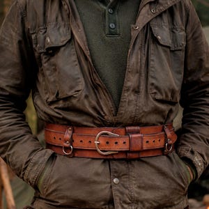 Aged brown bushcraft leather belt worn over an outdoor jacket in a forest camp setting, showing the belt as part of a rugged bushcraft and post apocalyptic outfit. Handmade wide utility belt with brass hardware, adjustable inner strap and side attachment rings for modular gear. Suitable for hiking gear, camping gear, tactical costume, military inspired outfit, steampunk belt, LARP armor, medieval ranger costume and Renaissance Faire clothing.