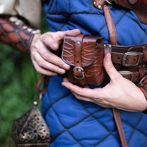 Può includere: Una borsa in pelle marrone con un motivo in rilievo, fissata con una fibbia. La borsa fa parte di un costume in stile medievale, indossato sopra un gilet trapuntato blu. Sono visibili anche una cintura e dei bracciali in pelle abbinati.