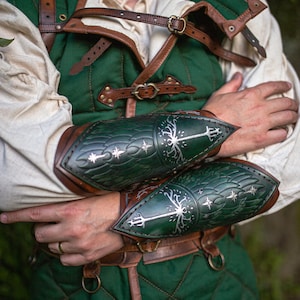 May include: Green and brown medieval armor set. The set includes a quilted vest, leather straps, and arm guards with a tree and star design. The arm guards are dark green with silver accents. The Outcast Props logo is visible.