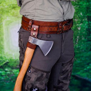 May include: A brown leather belt featuring a metal loop attachment designed to hold a hatchet. The belt has a wide, multi-layered construction with brass-colored rivets and a metal buckle. The hatchet has a wooden handle and a silver metal blade.