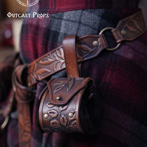 A brown floral embossed leather coin pouch hangs from a matching carved belt worn over a red and grey plaid skirt. The botanical design and aged leather finish give it a natural elven or druidic style. Perfect for renaissance faire or renfaire outfits, the pouch offers a soft suede body for carrying coins, dice or tiny essentials. A practical accessory for witches, herbalists, nobles or fantasy LARP characters seeking detailed leather gear.