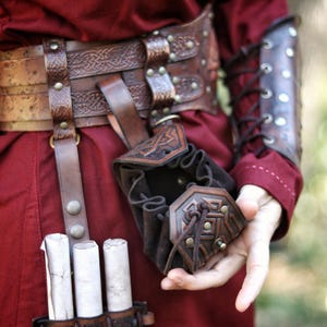 Handcrafted leather coin pouch worn on a belt, shown on a person dressed in fantasy medieval clothing. Small embossed leather pouch with Celtic geometric patterns, drawstring closure, and aged brown finish. Designed to carry coins, dice, keys, or small valuables. Suitable for Viking, Celtic, or dwarven characters, LARP, Renaissance fairs, cosplay, or everyday fantasy-inspired wear. Made from vegetable-tanned leather with artisanal aging.