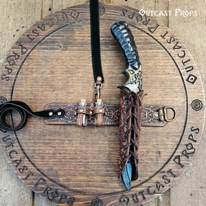 Könnte beinhalten: Ein handgefertigter Fantasy-Dolch mit detailliertem Griff und Lederscheide. Der Dolch wird auf einer Holzplakette mit dem Text "Outcast Props" präsentiert und enthält einen Lederriemen mit kleinen Fläschchen.