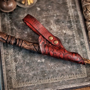 A red embossed leather wand holster rests on a weathered metal covered book while holding a carved wand. The holster features ornate patterns, brass toned rivets and a vivid hand dyed finish. The background includes sage bundles and fantasy props, creating an atmosphere suited for wizard cosplay, witch costume accessories, renaissance faire outfits and larp wand holder gear. The close framing highlights craftsmanship and texture.