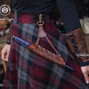 May include: A brown leather wand holder with embossed floral designs and brass studs, attached to a belt loop. The holder is paired with a matching leather arm cuff with leaf patterns and lace-up detailing. The image also shows a plaid skirt.