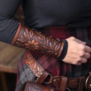 Side view of an aged brown floral leather bracer worn over a dark sleeve, paired with a renaissance faire belt bearing the same embossed leaf motif. The floral arm guard features hand tooled petals and natural shading that emphasize the artisan quality. This larp bracer functions as a fantasy cosplay accessory suited for elves, herbalists or fae characters, blending beautifully into medieval inspired outfits. Warm lighting highlights the engraved botanical pattern. Fae costume, ren faire, druid