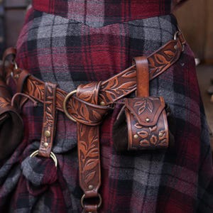 A full brown leather set with embossed botanical patterns is displayed on a person wearing a plaid skirt. The belt carries two accessories: a hipbag and a floral coin pouch. Brass rings and rivets complement the aged leather look. This renaissance faire inspired ensemble suits elves, witches, herbalists and nobles. The coin pouch works as a fantasy dice pouch or coin bag, offering practical storage for LARP and immersive costume wear.