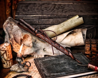May include: A brown leather-wrapped wooden writing quill with intricate carvings. The quill is resting on a vintage book with a skull and candle in the background.