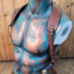 May include: A brown leather holster with adjustable straps for carrying a knife. The holster is worn on a mannequin with a blue and copper painted torso.