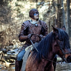 May include: A person wearing a leather armor and blue face paint rides a brown horse through a shallow stream in a forest.