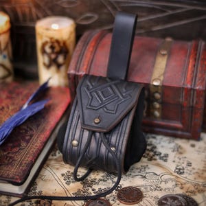 Black leather coin pouch with embossed geometric pattern and flap closure, displayed on a wooden surface with a chest, candle and illustrated map. The pouch is handmade from vegetable-tanned leather with careful aging, brass hardware and a soft suede body that expands for coins. A rear belt loop allows comfortable wear during events. This timeless leather coin pouch fits medieval, Viking, Celtic and fantasy styles, making it ideal for LARP, Renaissance fairs,