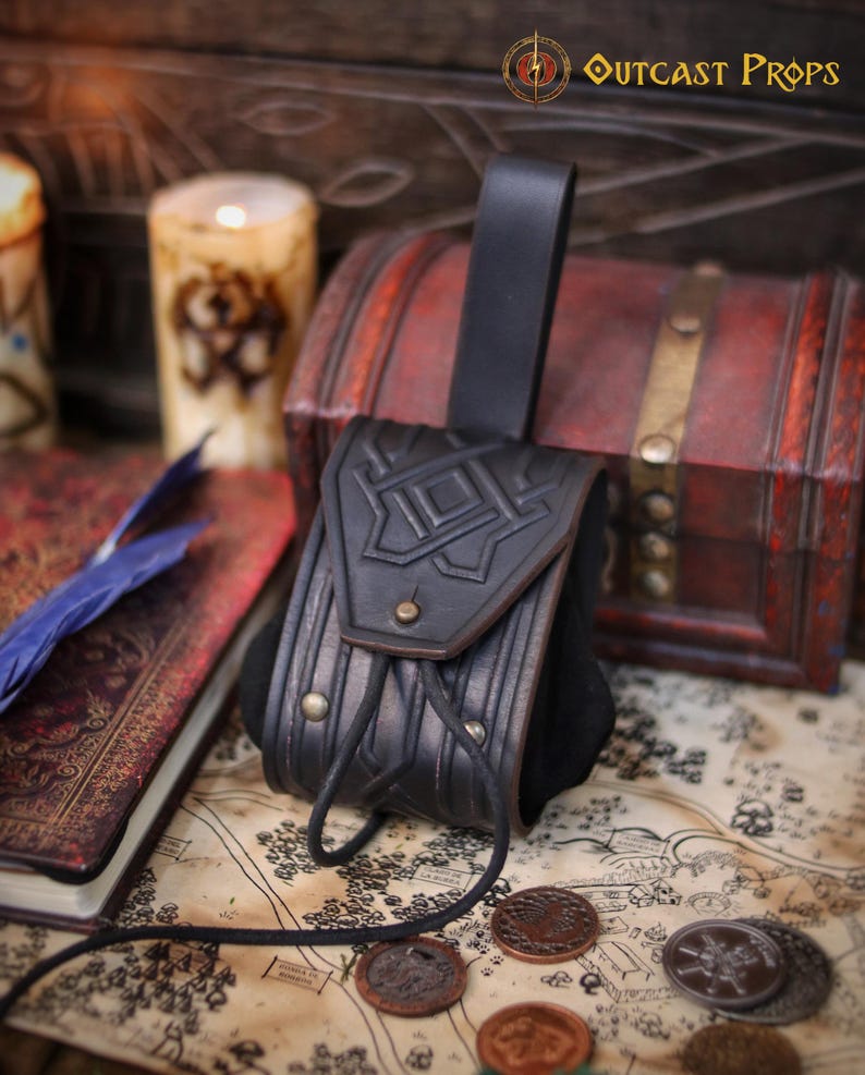 Black leather coin pouch with embossed geometric pattern and flap closure, displayed on a wooden surface with a chest, candle and illustrated map. The pouch is handmade from vegetable-tanned leather with careful aging, brass hardware and a soft suede body that expands for coins. A rear belt loop allows comfortable wear during events. This timeless leather coin pouch fits medieval, Viking, Celtic and fantasy styles, making it ideal for LARP, Renaissance fairs,