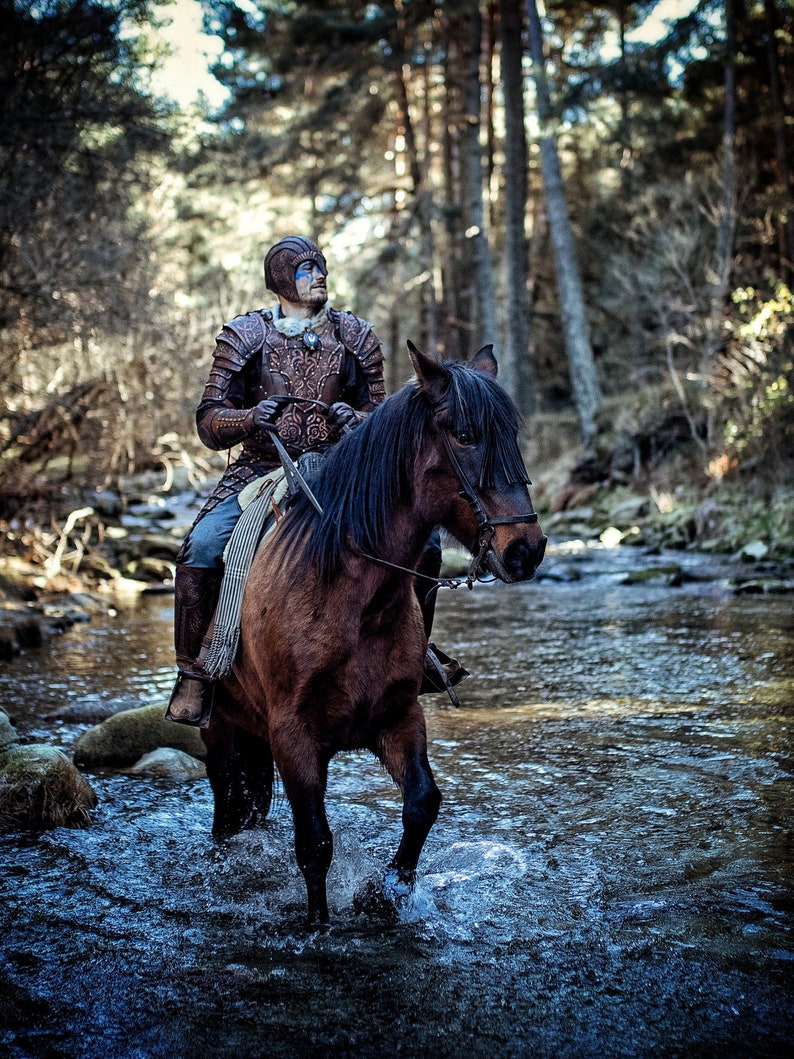 May include: A person wearing a leather armor and blue face paint rides a brown horse through a shallow stream in a forest.