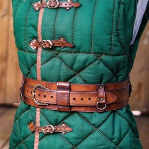 Aged brown bushcraft leather belt worn over a green quilted medieval gambeson, showing the wide leather frame, brass buckle, adjustable inner strap and side attachment rings. Handmade utility belt for LARP armor, medieval ranger costumes, Renaissance Faire outfits, bushcraft gear, hiking, camping, tactical costume, military inspired clothing, steampunk characters and fantasy adventurers. Built from premium vegetable tanned leather with aged finish and handcrafted details.