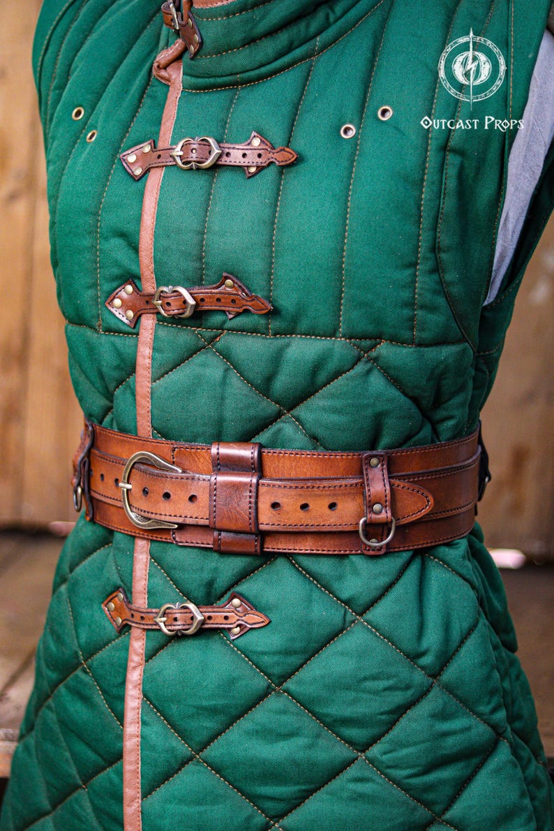 Aged brown bushcraft leather belt worn over a green quilted medieval gambeson, showing the wide leather frame, brass buckle, adjustable inner strap and side attachment rings. Handmade utility belt for LARP armor, medieval ranger costumes, Renaissance Faire outfits, bushcraft gear, hiking, camping, tactical costume, military inspired clothing, steampunk characters and fantasy adventurers. Built from premium vegetable tanned leather with aged finish and handcrafted details.
