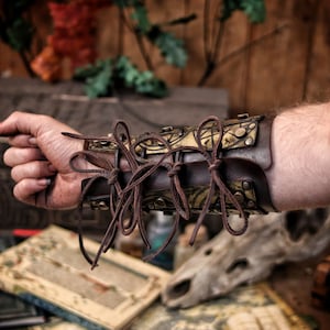 Golden fantasy bracers with matching armguard, engraved with magical runes and ritual symbols, shown in an arcane tabletop scene with scrolls and potions. Perfect for LARP witch armor, warlock costume or alchemist outfit. Handmade with aged leather and adjustable laces, these vambraces are a high-quality accessory for medieval, fantasy or occult cosplay enthusiasts.