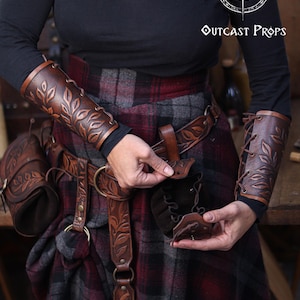 A person wearing brown embossed leather bracers and a matching belt demonstrates how to open a floral leather coin pouch. The plaid skirt and rustic workshop background create a renaissance faire atmosphere. The pouch’s suede interior and drawstring are visible as it is held open. This handcrafted renfaire pouch suits elves, witches, herbalists and nobles, working as a leather coin bag or fantasy dice pouch for immersive LARP and cosplay costumes.