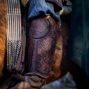 May include: Close-up of a brown leather leg armor with intricate designs. The armor is worn over blue pants and has straps and buckles.