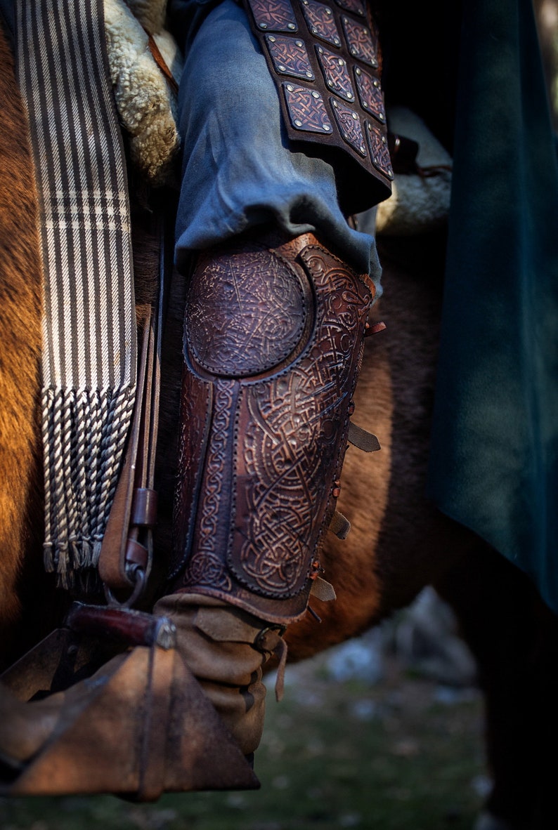 May include: Close-up of a brown leather leg armor with intricate designs. The armor is worn over blue pants and has straps and buckles.