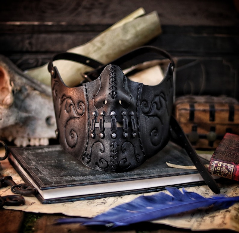 A black leather mask with a skull design. The mask has silver rivets and stitching. The mask is on a book with a blue feather on top of it. Death Wizard Leather Mask M2 - Handcrafted Mask for Dark mages witches and powerful sorcerers with Intricate Details and Metallic Finish
