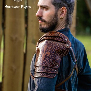 May include: A brown leather shoulder pauldron with detailed embossed patterns, featuring a fleur-de-lis. The pauldron is worn over a dark blue shirt. The Outcast Props logo is visible in the upper left corner.