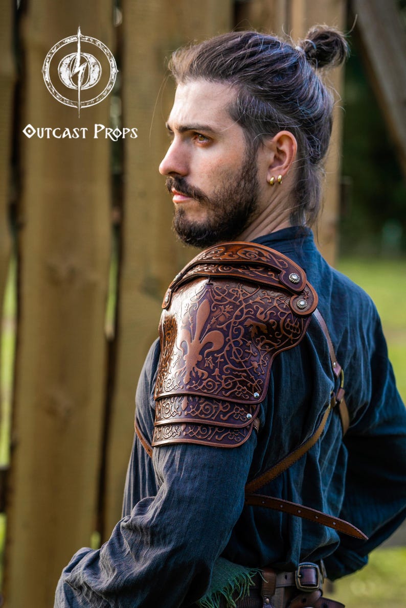May include: A brown leather shoulder pauldron with detailed embossed patterns, featuring a fleur-de-lis. The pauldron is worn over a dark blue shirt. The Outcast Props logo is visible in the upper left corner.