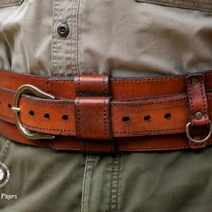 May include: A wide, brown leather belt featuring a brass-toned buckle, multiple adjustment holes, and integrated leather loops. The belt includes a small metal D-ring attached to the side with a riveted leather strap. The text Outcast Props is visible.