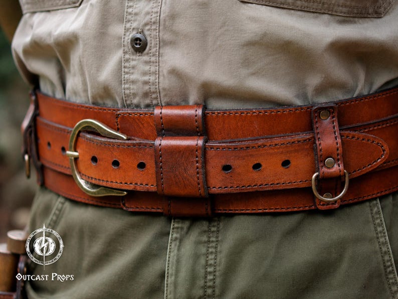 May include: A wide, brown leather belt featuring a brass-toned buckle, multiple adjustment holes, and integrated leather loops. The belt includes a small metal D-ring attached to the side with a riveted leather strap. The text Outcast Props is visible.