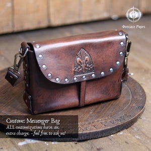 May include: A brown leather messenger bag with a flap closure, metal studs, and a decorative emblem. The bag has a shoulder strap and metal hardware. The text "Custom: Messenger Bag" is visible.