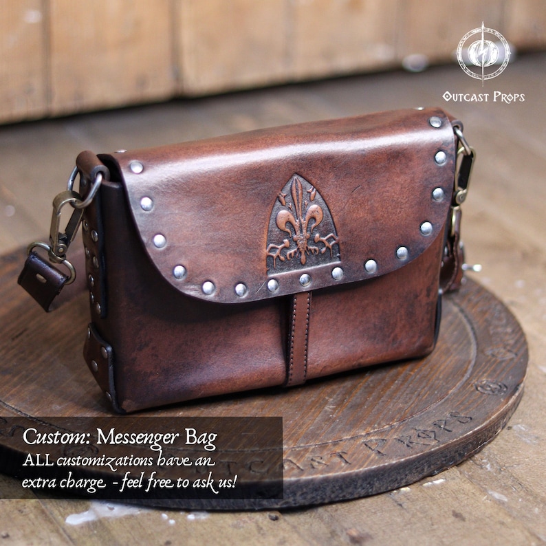 May include: A brown leather messenger bag with a flap closure, metal studs, and a decorative emblem. The bag has a shoulder strap and metal hardware. The text "Custom: Messenger Bag" is visible.