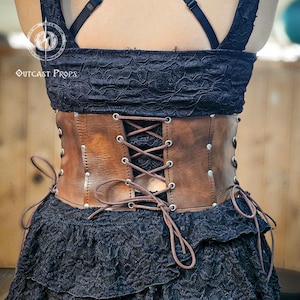Back view of the aged brown witch leather corset featuring symmetrical lacing for adjustable fit. Made from full-grain vegetable-tanned leather with nickel rivets, this underbust corset enhances gothic and steampunk outfits. Ideal for ren faire costumes or fantasy festivals, it combines historical design and artisan detail. Every Outcast Props corset is hand-aged, making each piece unique in tone and texture.