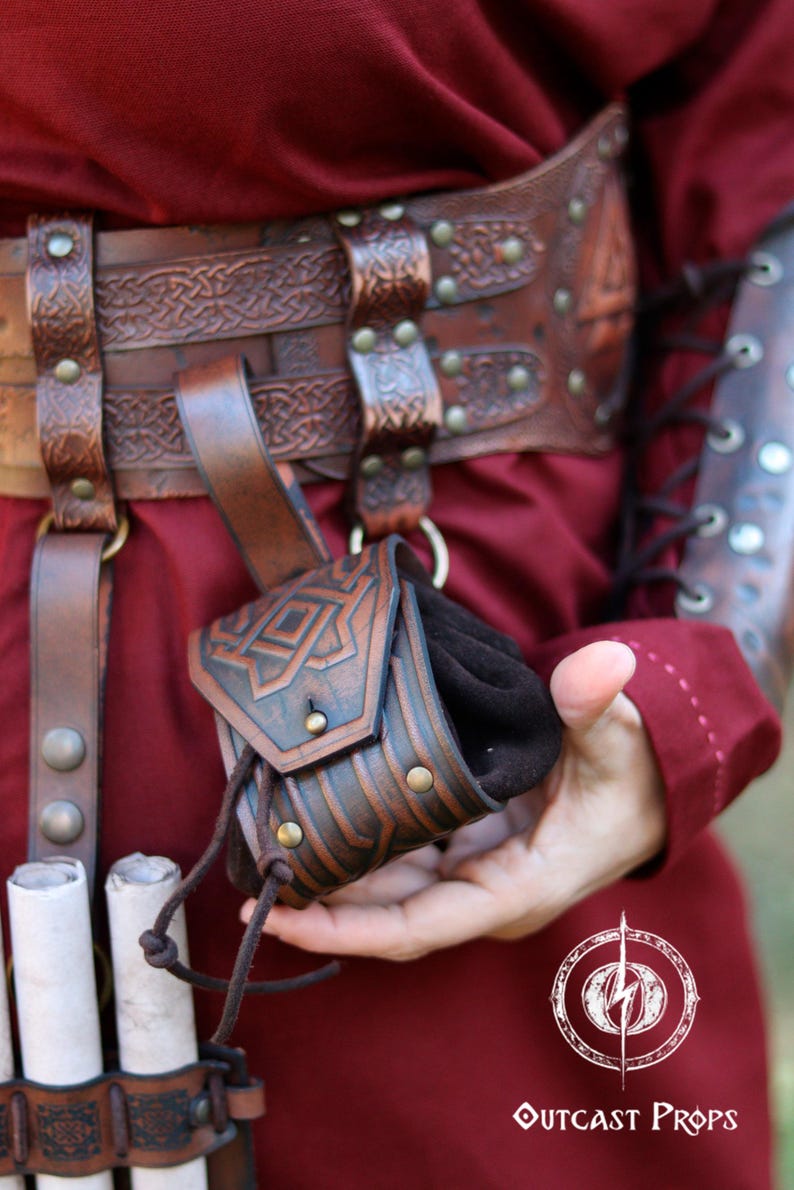 Close-up of a small decorated leather coin pouch held in hand, showing its size and texture. Handcrafted pouch with embossed Celtic geometric designs, aged brown finish, and drawstring closure. Ideal for carrying coins, dice, or keys. A compact leather accessory for Viking, Celtic, or dwarven aesthetics, suitable for LARP, Renaissance fairs, cosplay, or as a small handmade leather gift.