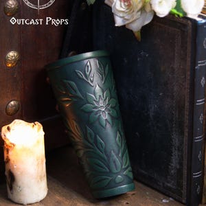 A single aged green floral leather bracer leaning against antique books with white roses above it. This renaissance faire bracer features deep embossed botanical patterns and artisan aging, ideal as an elf cosplay accessory, fae costume accessory or larp bracer. Warm candlelight enhances its floral arm guard texture and handcrafted look, perfect for romantic fantasy or woodland themed outfits. Fae costume, ren faire, druid larp, leaf design, floral design.