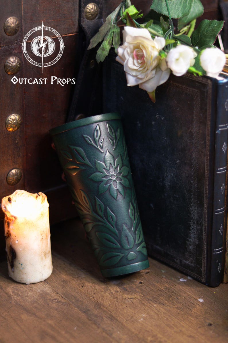 A single aged green floral leather bracer leaning against antique books with white roses above it. This renaissance faire bracer features deep embossed botanical patterns and artisan aging, ideal as an elf cosplay accessory, fae costume accessory or larp bracer. Warm candlelight enhances its floral arm guard texture and handcrafted look, perfect for romantic fantasy or woodland themed outfits. Fae costume, ren faire, druid larp, leaf design, floral design.