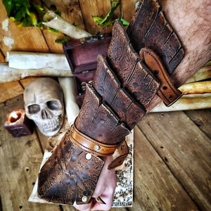 A brown handmade Dragon Scales Leather Bracer is worn on a man’s forearm, fully fastened and resting over a vintage map surrounded by fantasy props like a skull and candle. The layered leather plates resemble dragon hide, with an aged finish and rugged style. Perfect for LARP warriors, monster hunters or medieval cosplay. The leather is vegetable-tanned and hand-painted using Outcast Props’ signature weathering technique.