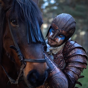 Viking warrior in full handcrafted Celtic leather armor Sedrik, detailed with Nordic knot engravings and fantasy Viking leather shoulder pauldrons, photographed outdoors petting a brown horse. Helmet with engraved leather and face paint complete the Nordic LARP armor look. Ideal for Viking cosplay, fantasy LARP armor, Norse warrior costume, Viking leather armor set, horse-mounted warrior outfit, Nordic reenactment, and Celtic fantasy costume. Premium leather craftsmanship and textures.