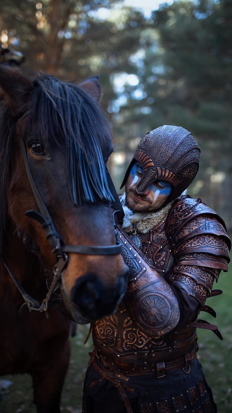 Viking warrior in full handcrafted Celtic leather armor Sedrik, detailed with Nordic knot engravings and fantasy Viking leather shoulder pauldrons, photographed outdoors petting a brown horse. Helmet with engraved leather and face paint complete the Nordic LARP armor look. Ideal for Viking cosplay, fantasy LARP armor, Norse warrior costume, Viking leather armor set, horse-mounted warrior outfit, Nordic reenactment, and Celtic fantasy costume. Premium leather craftsmanship and textures.