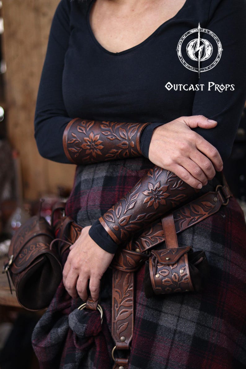 A pair of aged brown floral leather bracers is shown worn on both arms while the wearer stands in a relaxed pose with crossed arms. The renaissance faire bracer pattern displays embossed leaves and a central flower that highlight the hand dyed finish. Below, a matching floral belt and pouch complete the look, creating an elegant floral arm guard ensemble ideal as an elf cosplay accessory or larp bracer set. Perfect for fantasy themed costumes and woodland characters. Fae costume, ren faire druid