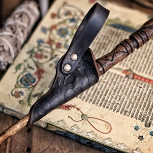 Close up of a black embossed leather wand holster holding a carved wooden wand, resting on an illuminated medieval manuscript decorated with floral borders. The holster loop shows two rounded rivets and the leather texture appears aged and shaped by hand. Perfect accessory for wizard cosplay, witch costume, fantasy mages and larp wand holder gear. The scene includes bundled sage and old books, reinforcing the magical, alchemist inspired atmosphere.